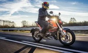 Motocyklista w kasku i kurtce jedzie pomarańczowym motocyklem po drodze. Słońce przebija się przez chmury, tworząc refleksy. To jak odpalic motor i ruszyć w drogę!
