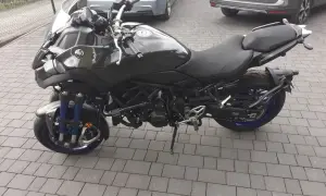 Czarny motocykl Yamaha Niken kat b. na kostce brukowej, z niebieskimi elementami zawieszenia i felg.