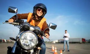 Uśmiechnięta kursantka na motocyklu, gotowa na swój pierwszy kurs na motor. Instruktor obserwuje z tyłu.