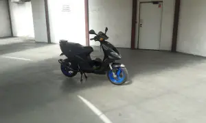 Czarny Piaggio NRG MC3 z niebieskimi felgami stoi na betonowej posadzce w garażu. Obok leży zielony kask.