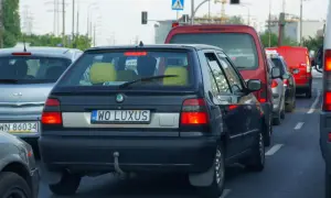 Czarny samochód z indywidualnymi tablicami rejestracyjnymi "WO LUXUS" w korku. Obok inne auta, w tym czerwony van i biała furgonetka.