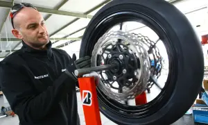 Mechanik w rękawiczkach dokonuje wymiany opon motocyklowych. Koło z tarczami hamulcowymi jest zamocowane na czerwonym stojaku.