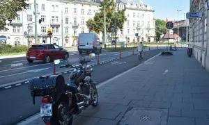 Motocykl zaparkowany na chodniku, obok ścieżki rowerowej. W tle samochody i budynki.