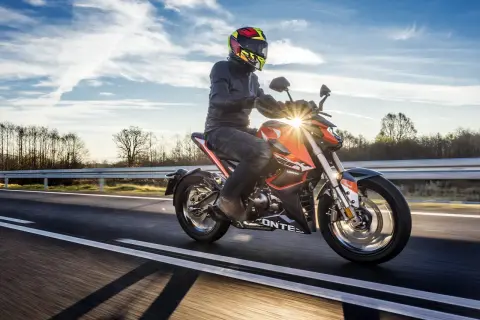 Motocyklista w kasku i kurtce jedzie pomarańczowym motocyklem po drodze. Słońce przebija się przez chmury, tworząc refleksy. To jak odpalic motor i ruszyć w drogę!