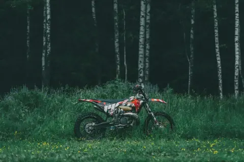Motocykl cross w terenie, zaparkowany wśród wysokiej trawy i dzikich kwiatów, na tle mrocznego lasu.