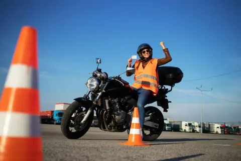 Szczęśliwa motocyklistka z radością prezentuje swoje nowe prawo jazdy kat A, siedząc na motorze na placu manewrowym.