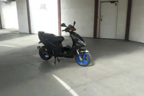 Czarny Piaggio NRG MC3 z niebieskimi felgami stoi na betonowej posadzce w garażu. Obok leży zielony kask.