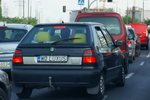 Czarny samochód z indywidualnymi tablicami rejestracyjnymi "WO LUXUS" w korku. Obok inne auta, w tym czerwony van i biała furgonetka.