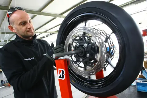 Mechanik w rękawiczkach dokonuje wymiany opon motocyklowych. Koło z tarczami hamulcowymi jest zamocowane na czerwonym stojaku.