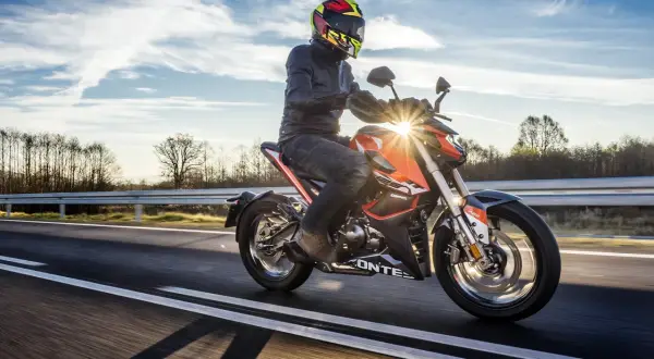 Motocyklista w kasku i kurtce jedzie pomarańczowym motocyklem po drodze. Słońce przebija się przez chmury, tworząc refleksy. To jak odpalic motor i ruszyć w drogę!