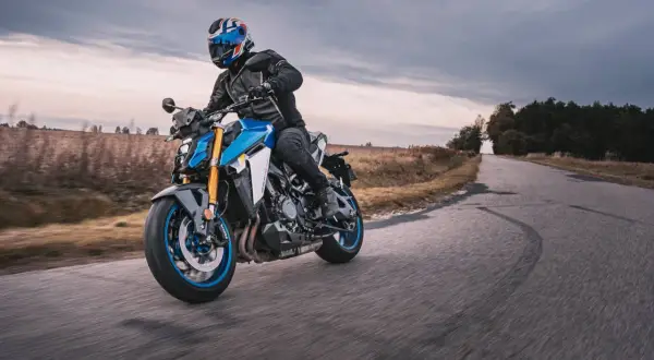 Motocyklista w kasku jedzie niebieskim motocyklem po drodze. W tle pola i las, chmury na niebie.