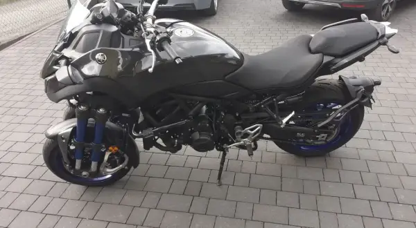 Czarny motocykl Yamaha Niken kat b. na kostce brukowej, z niebieskimi elementami zawieszenia i felg.