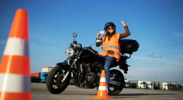 Szczęśliwa motocyklistka z radością prezentuje swoje nowe prawo jazdy kat A, siedząc na motorze na placu manewrowym.