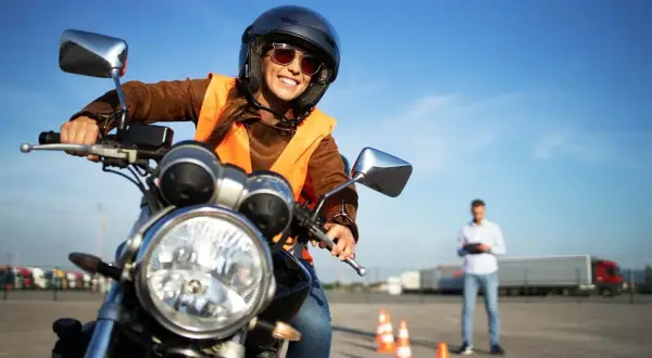 Uśmiechnięta kursantka na motocyklu, gotowa na swój pierwszy kurs na motor. Instruktor obserwuje z tyłu.