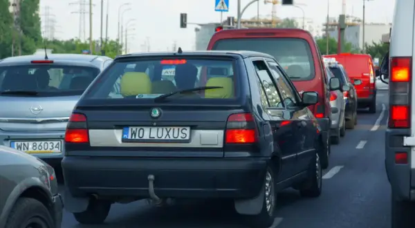Czarny samochód z indywidualnymi tablicami rejestracyjnymi "WO LUXUS" w korku. Obok inne auta, w tym czerwony van i biała furgonetka.