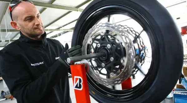 Mechanik w rękawiczkach dokonuje wymiany opon motocyklowych. Koło z tarczami hamulcowymi jest zamocowane na czerwonym stojaku.