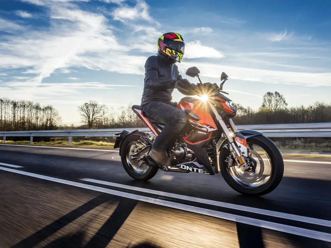 Motocyklista w kasku i kurtce jedzie pomarańczowym motocyklem po drodze. Słońce przebija się przez chmury, tworząc refleksy. To jak odpalic motor i ruszyć w drogę!