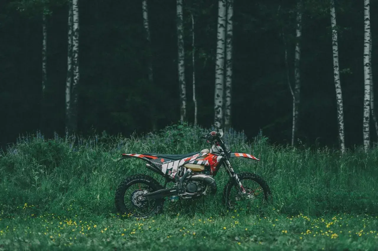 Motocykl cross w terenie, zaparkowany wśród wysokiej trawy i dzikich kwiatów, na tle mrocznego lasu.