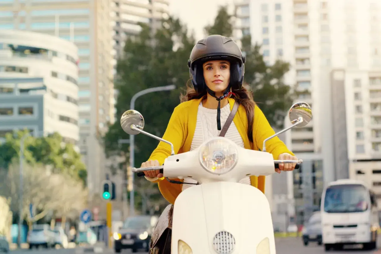 Kobieta w żółtym swetrze i kasku jedzie skuterem po mieście. Czy na skuter trzeba mieć prawo jazdy? To pytanie zadaje sobie wiele osób.