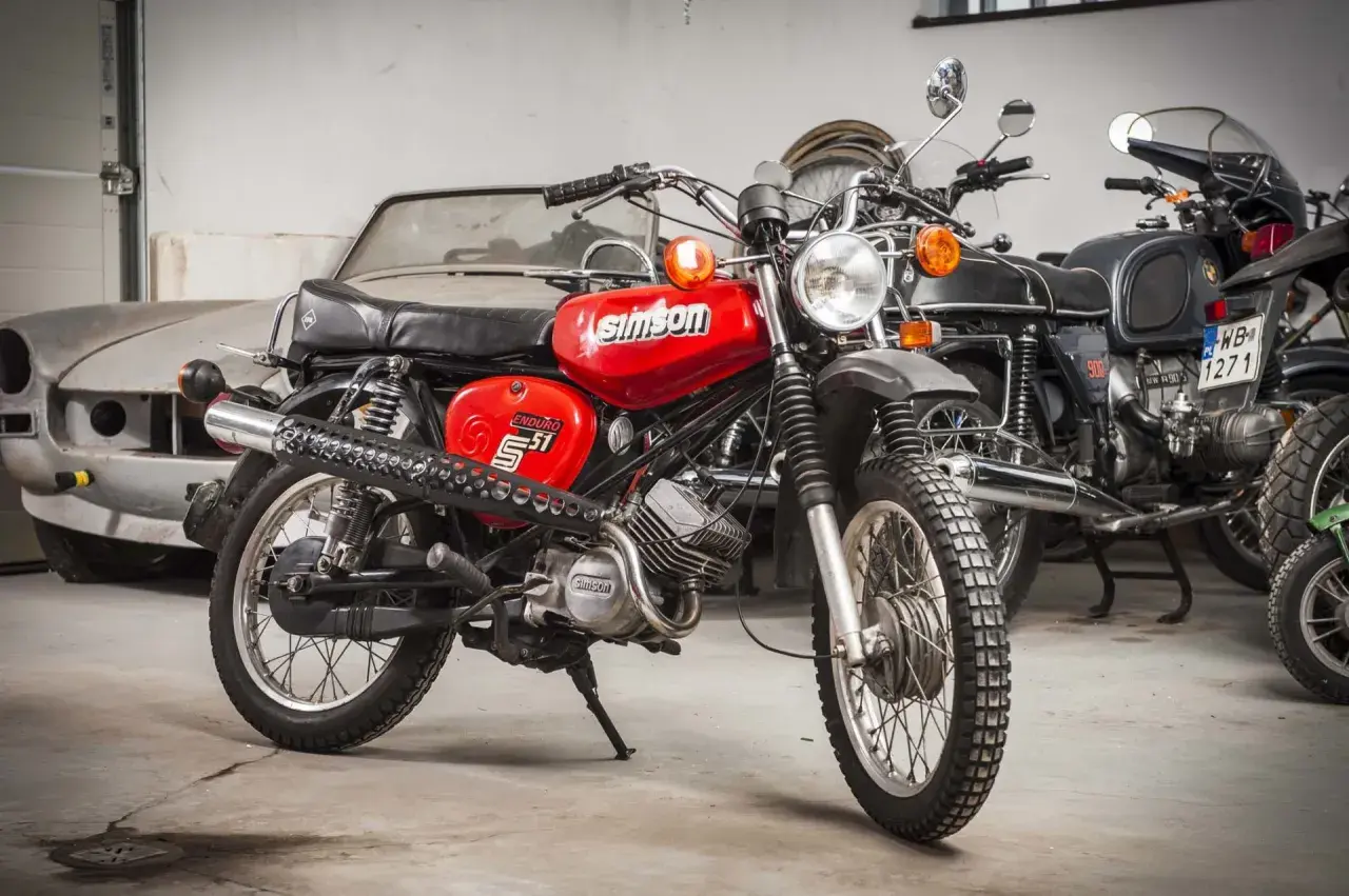 Czerwony Simson S51 Enduro, z widocznymi danymi technicznymi na baku. Obok inne motocykle i stary samochód.