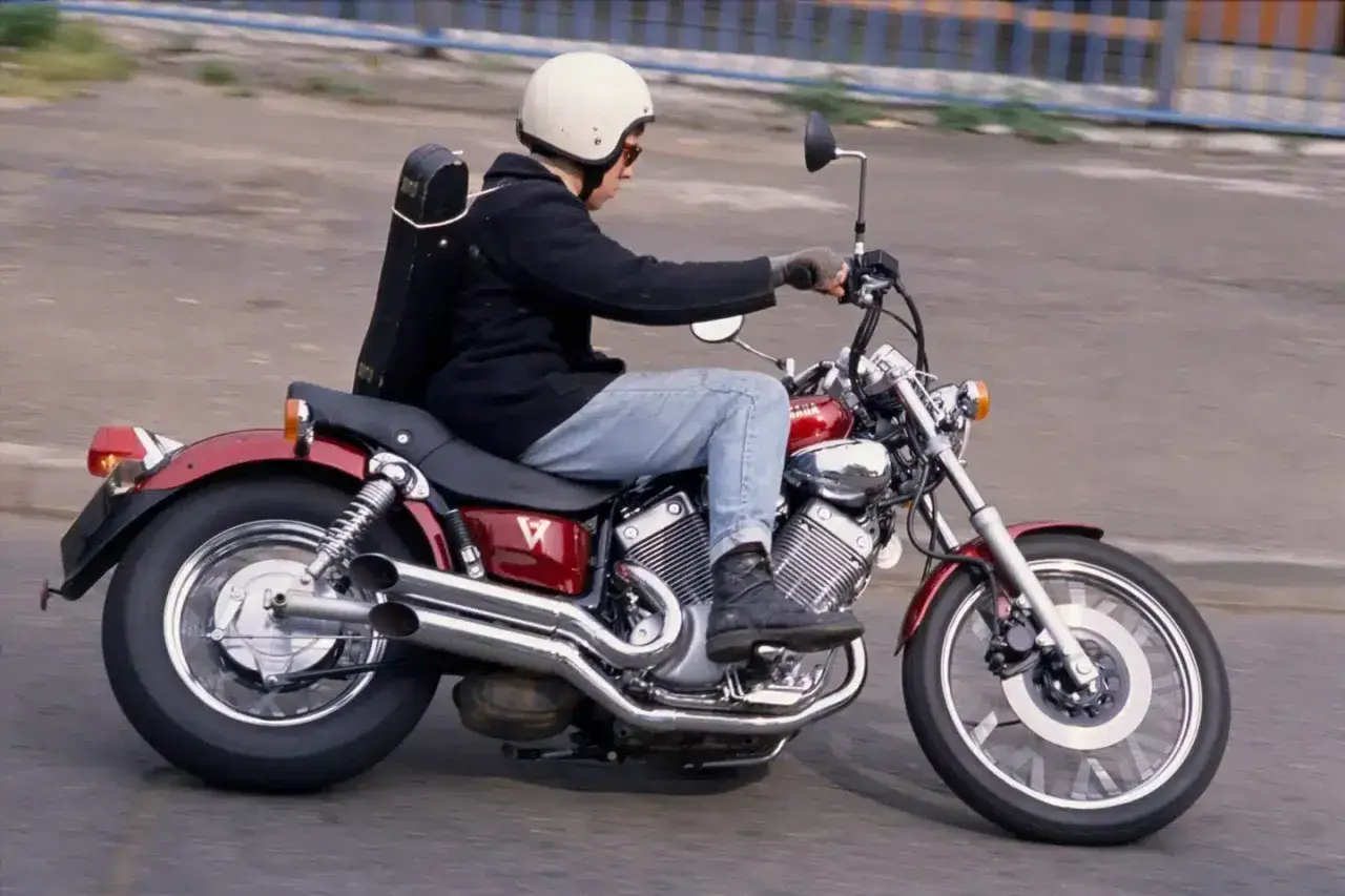 Mężczyzna w kasku jedzie na czerwonym motocyklu Yamaha Virago, z futerałem na instrumenty muzyczne na plecach.