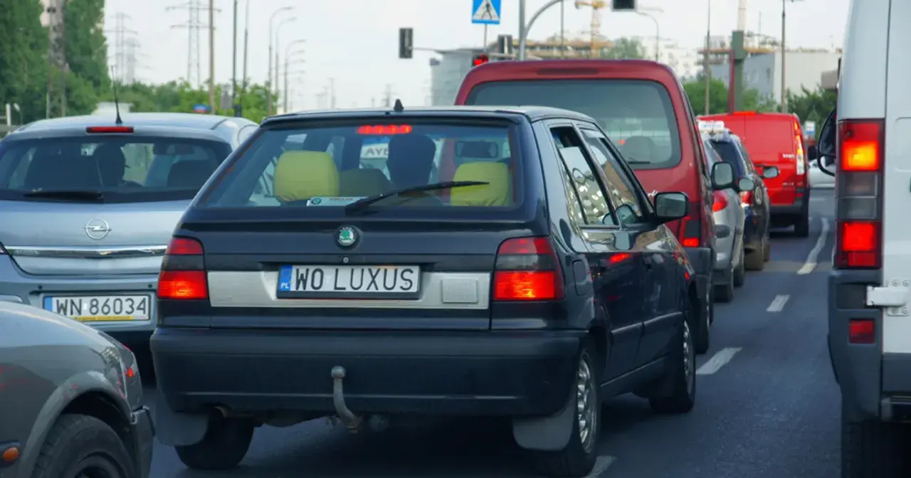 Czarny samochód z indywidualnymi tablicami rejestracyjnymi "WO LUXUS" w korku. Obok inne auta, w tym czerwony van i biała furgonetka.