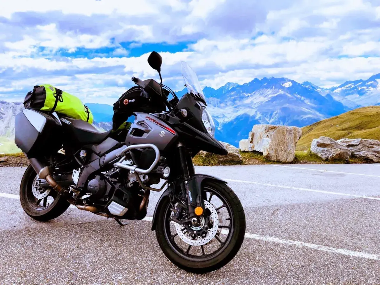 Gotowy na przygodę motocykl szosowy Suzuki V-Strom z sakwami i jaskrawym plecakiem czeka na tle majestatycznych gór i błękitnego nieba.