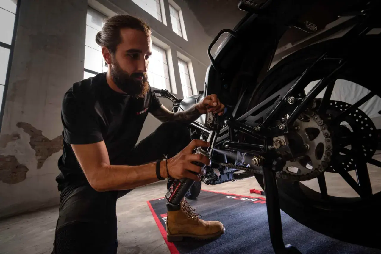 Mężczyzna z brodą konserwuje łańcuch motocykla, pokazując, jak założyć łańcuch.