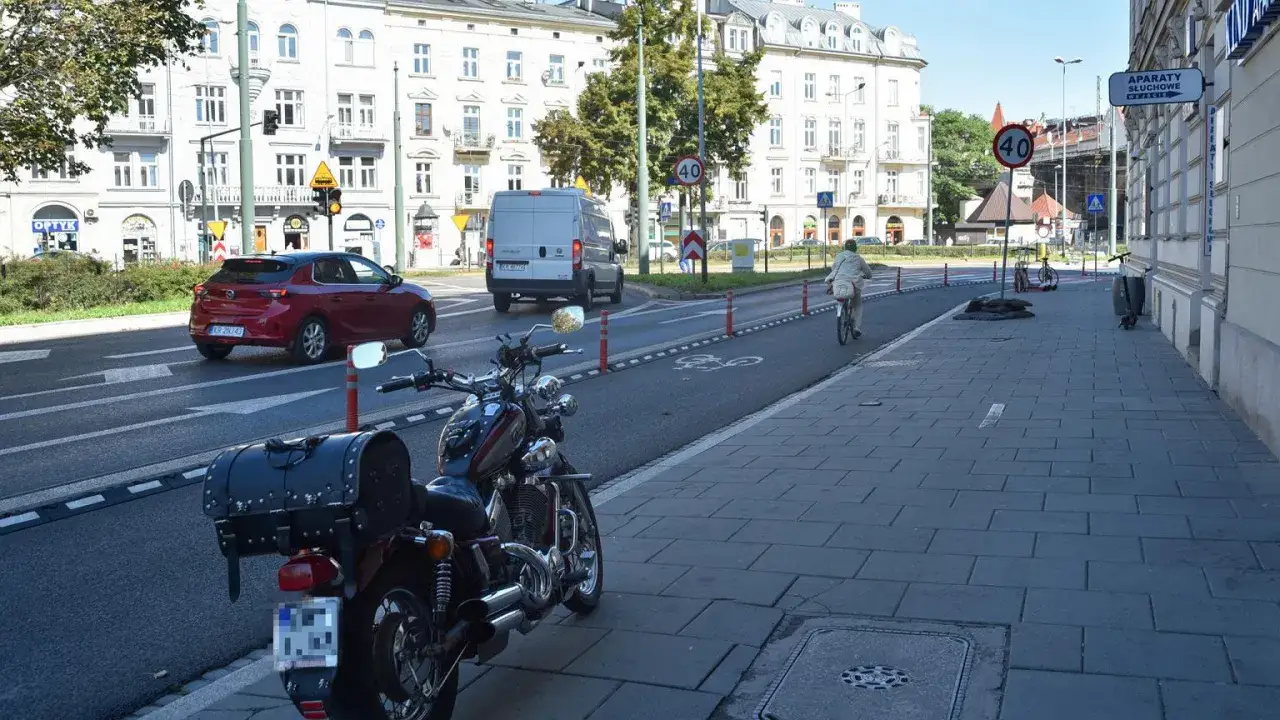Motocykl zaparkowany na chodniku obok ścieżki rowerowej. W tle czerwony samochód i biała furgonetka.