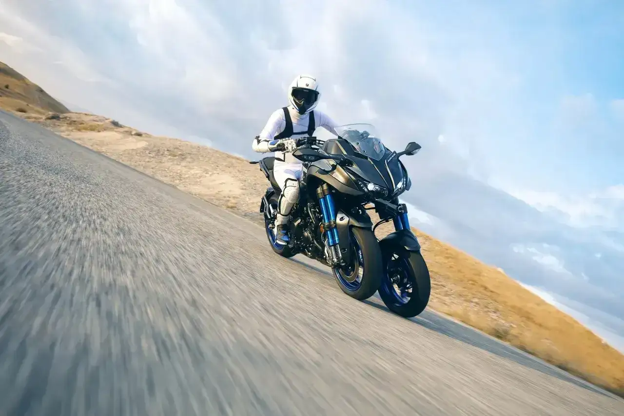 Motocyklista w białym kombinezonie na sportowym motocyklu Yamaha Niken kat b. pędzi po krętej drodze w g&oacute;rzystym terenie.
