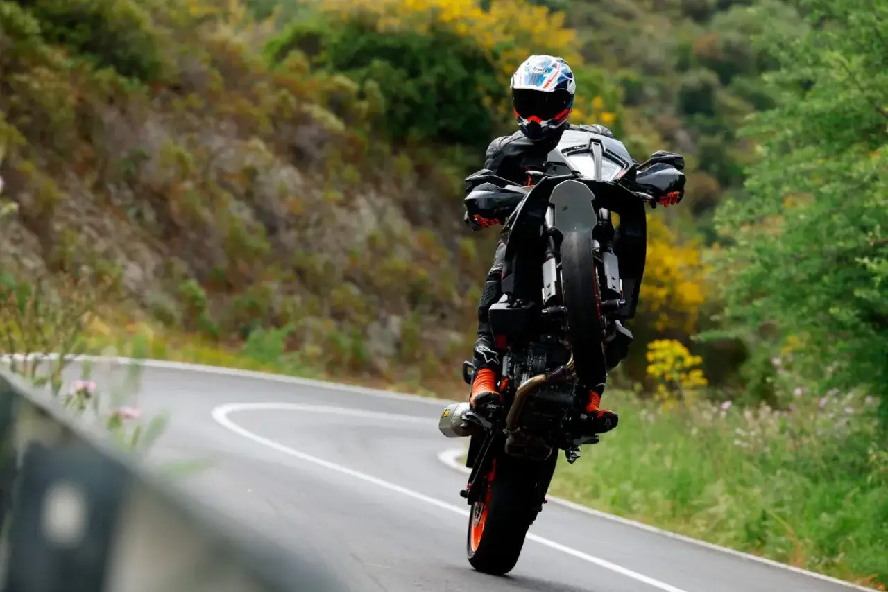 Motocyklista na KTM 890 SMT wykonuje wheelie na krętej drodze wśr&oacute;d zieleni i ż&oacute;łtych kwiat&oacute;w.