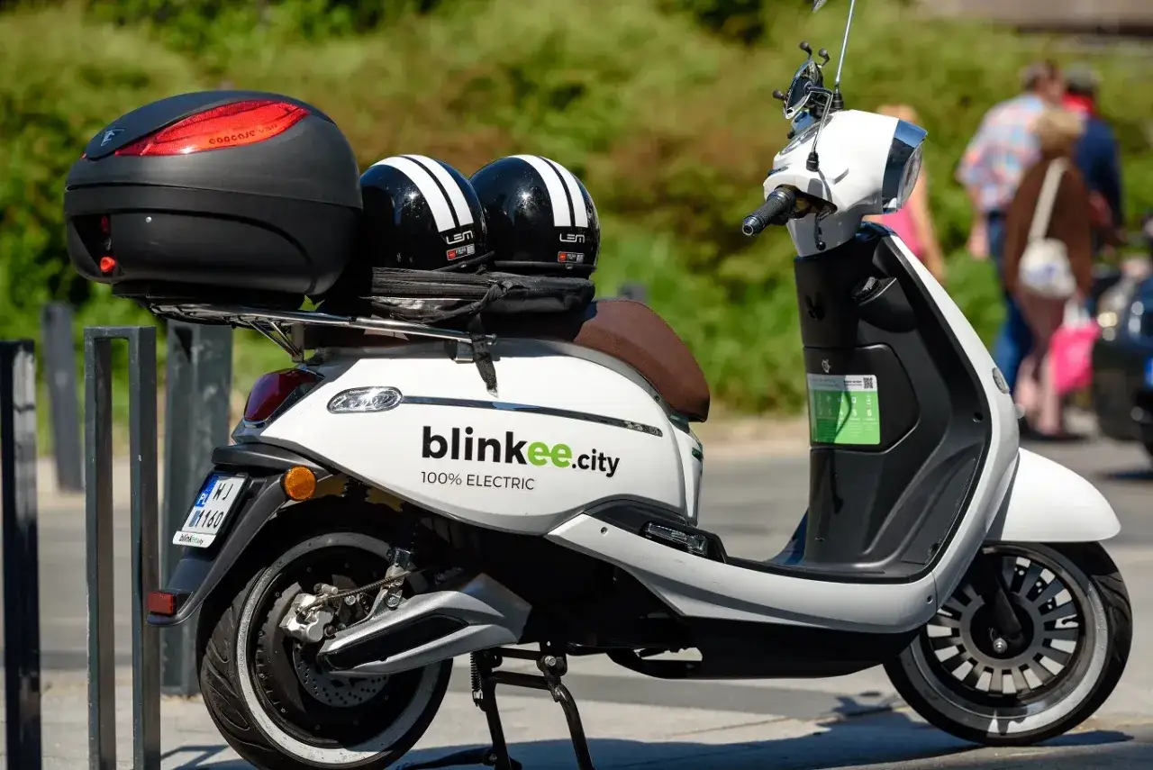 Biały skuter elektryczny Blinkee.city z dwoma kaskami na siedzeniu, idealny do miejskich podr&oacute;ży bez prawa jazdy.