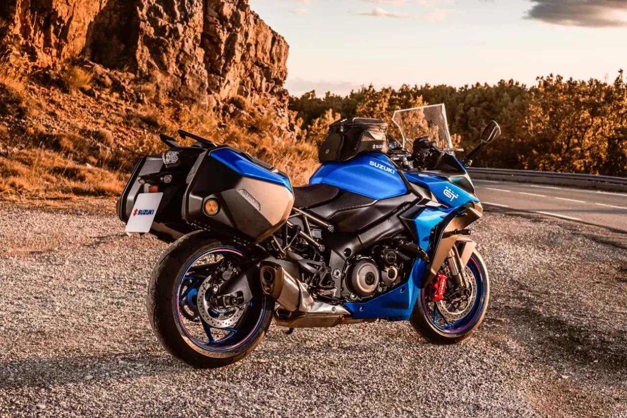 Niebieski motocykl Suzuki GSX S1000 GT z kuframi, zaparkowany na szutrowej drodze przy skalistym zboczu.