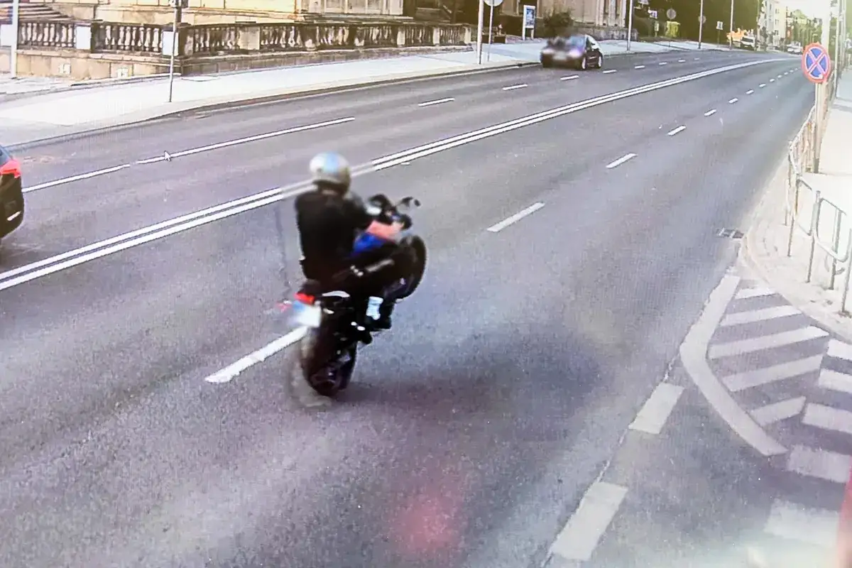 Motocyklista wykonuje jazdę na jednym kole, co grozi mandatem. Na drodze widać inne pojazdy i budynki.