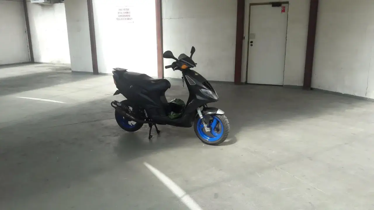 Czarny Piaggio NRG MC3 z niebieskimi felgami stoi na betonowej posadzce w garażu. Obok leży zielony kask.