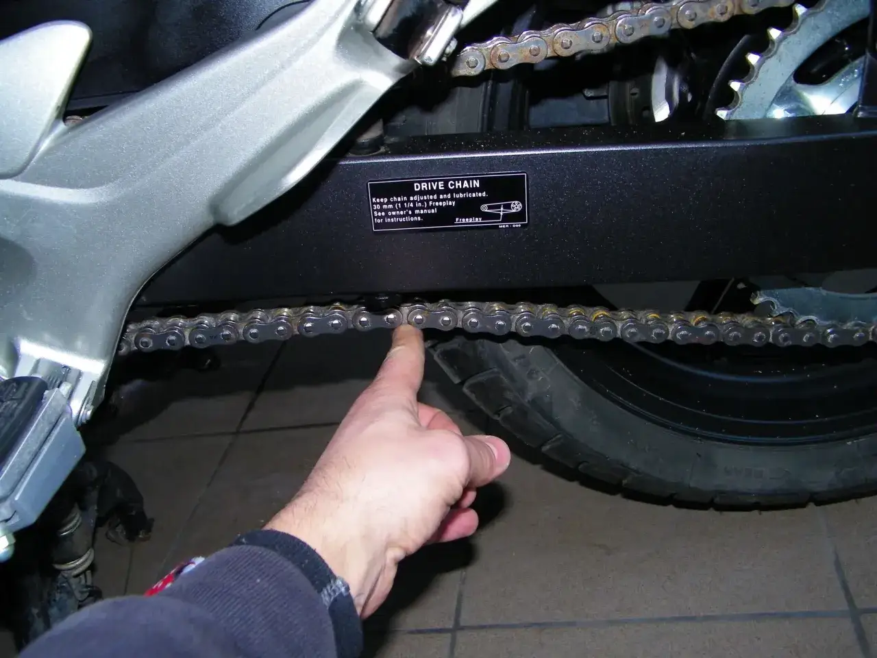 Sprawdzanie luzu łańcucha napędowego motocykla, kluczowe przy pytaniu jak założyć łańcuch. Wskaz&oacute;wka na tabliczce m&oacute;wi o 30 mm luzu.
