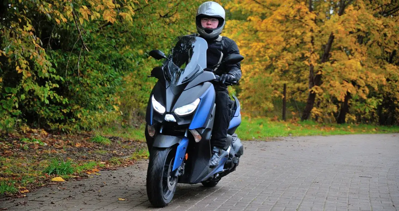Kierowca w kasku na skuterze Yamaha X-MAX 400, otoczony jesienną przyrodą. Czy osiągnie swoją prędkość maksymalną?