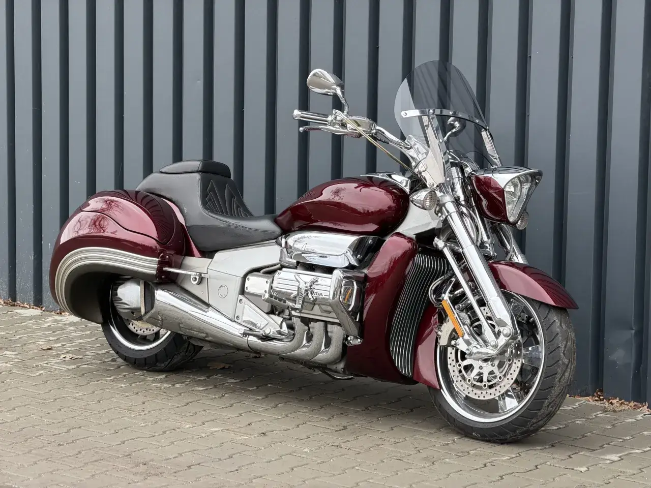 Lśniąca, wiśniowa Honda Valkyrie z chromowanymi detalami i dużą szybą, zaparkowana na chodniku przed szarą, pionową ścianą.
