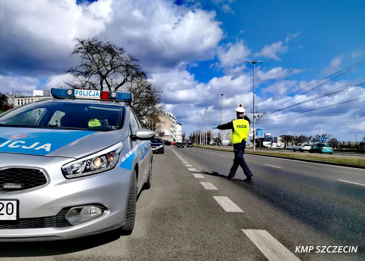 Policjant kieruje ruchem, kontrolując, jaka kara za jazdę bez przeglądu grozi kierowcom. Radiow&oacute;z stoi na poboczu.