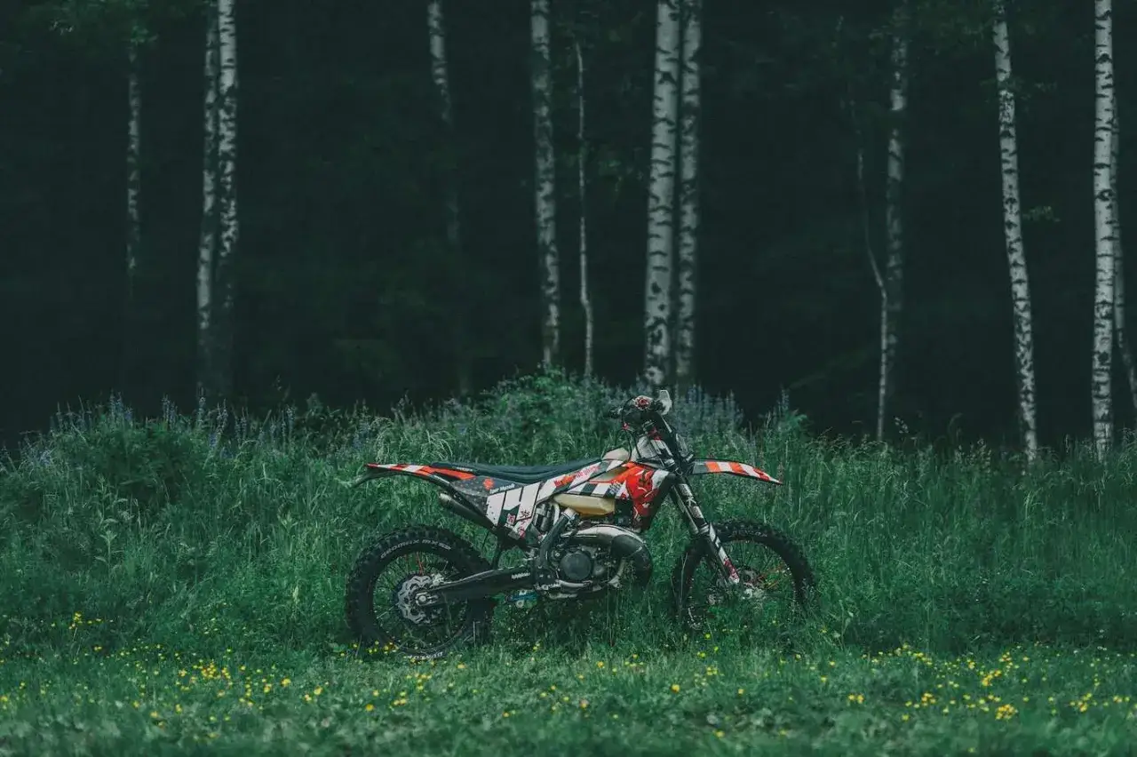 Motocykl crossowy w trawie, gotowy na przygodę. Idealne miejsce, gdzie można jeździć crossem, wśr&oacute;d drzew i dzikich kwiat&oacute;w.