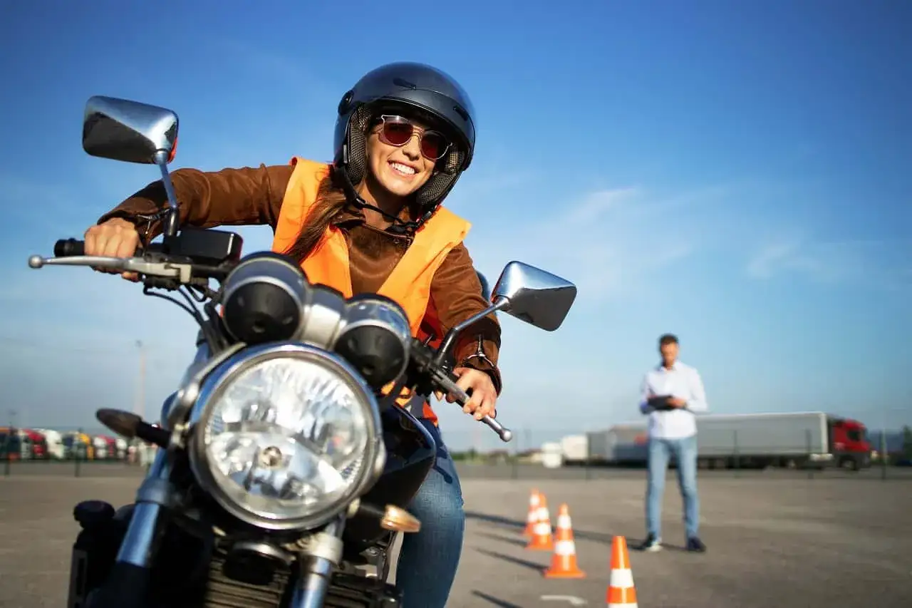 Uśmiechnięta kursantka na motocyklu, w kasku i kamizelce odblaskowej, uczy się jeździć. Zastanawiasz się, ile kosztują prawo jazdy na motor?