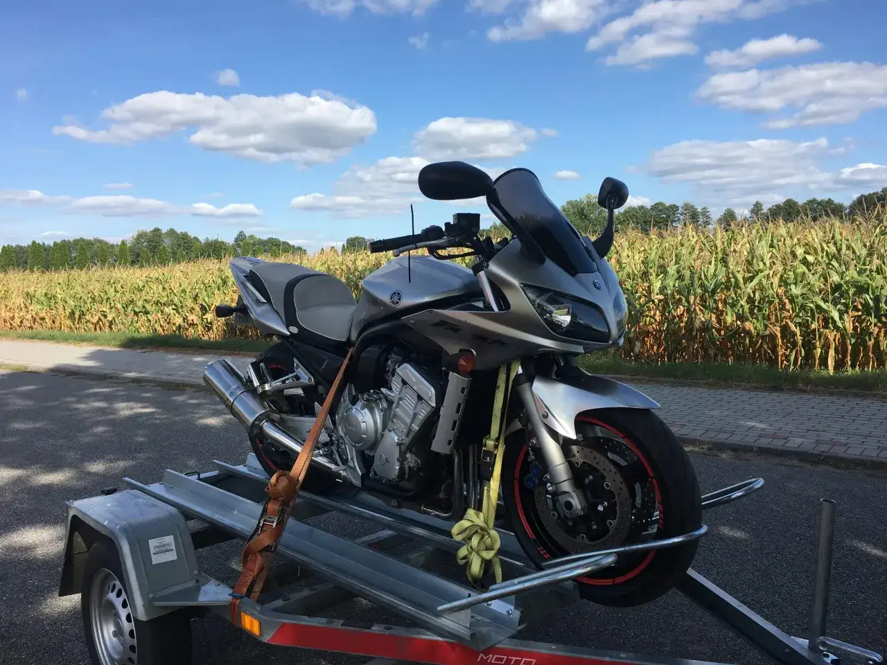 Srebrny motocykl Yamaha na przyczepce, gotowy do rejestracji motocykla z zagranicy. W tle pole kukurydzy i błękitne niebo.