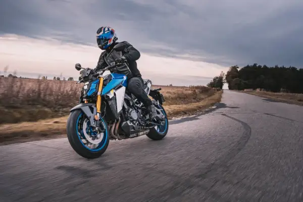 Motocyklista w kasku jedzie niebieskim motocyklem po drodze. W tle pola i las, chmury na niebie.