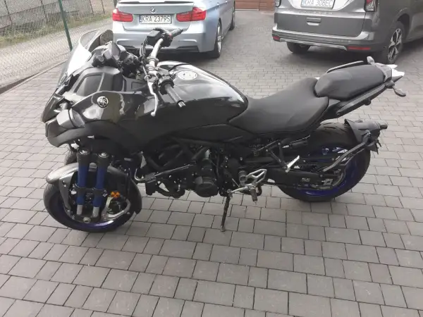 Czarny motocykl Yamaha Niken kat b. na kostce brukowej, z niebieskimi elementami zawieszenia i felg.