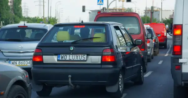 Czarny samochód z indywidualnymi tablicami rejestracyjnymi "WO LUXUS" w korku. Obok inne auta, w tym czerwony van i biała furgonetka.