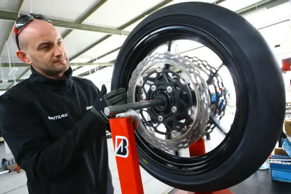 Mechanik w rękawiczkach dokonuje wymiany opon motocyklowych. Koło z tarczami hamulcowymi jest zamocowane na czerwonym stojaku.
